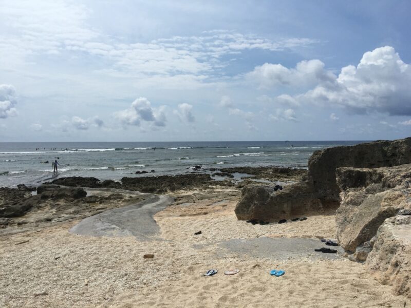 Rocks and a small patch of sand at the edge of the reef at Komesu Coast, with surfers in the water